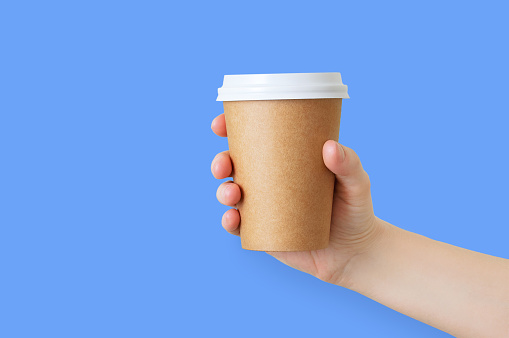 Paper brown cup of coffee in the hand of a caucasian girl. Blue background.