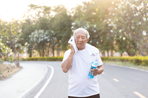Close up Asian elderly wiping sweat with towel after exercised running on road in public park that has sunshine in the evening. health lifestyle and exercise Concept.