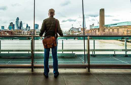 Wide angle color image depicting a rear view of a male commuter in his 50s or 60s looking through the window at a railway station in central London, overlooking the modern city skyline and river Thames. The man wears casual denim jeans, a green down jacket and a brown leather messenger bag.