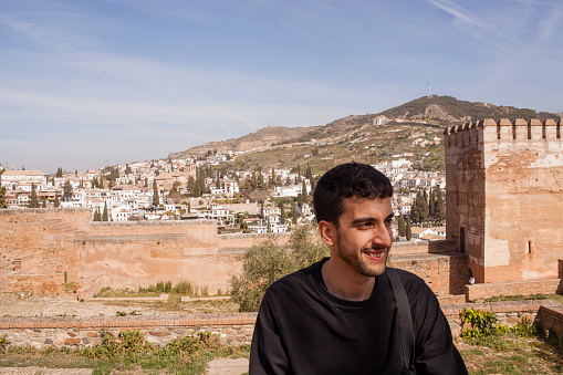 Adult Moroccan bearded man traveler smiling enjoying the views from Alhambra castle of Granada in Andalusia while resting on a old city landscape background on a sunny day. Happiness travelling Spain