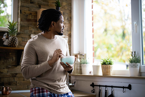 Young African American man day dreaming while looking through window during breakfast time in the kitchen.