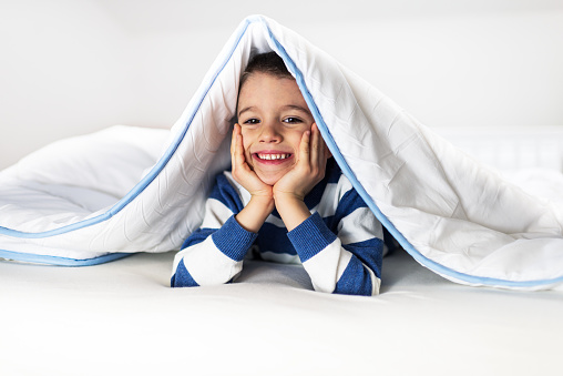 Beautiful boy is covered with blanket on bed, in bedroom. He is smiling.