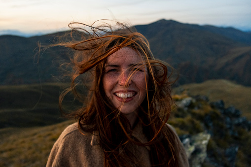 A portrait of a young caucasian woman looking at the camera and smiling while on holiday in New Zealand. The wind is blowing her hair over her face.