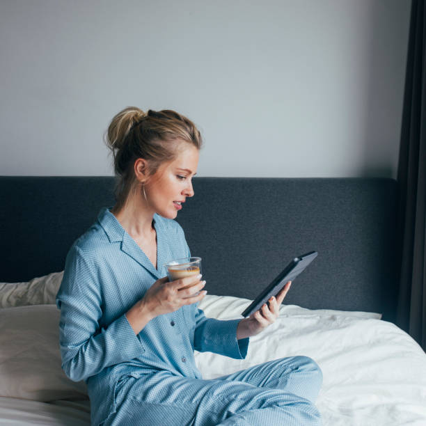 istock Morning Routine: Beautiful Blonde Woman in Pyjamas Sitting on the Bed,  Drinking Coffee and Reading from a Tablet 1211550607
