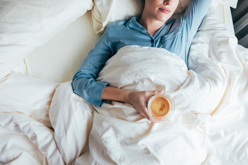 Cropped image of a Caucasian woman in blue pyjamas lying on the bed holding her morning latte in her hand.