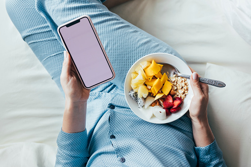Close up shot of a woman's hands holding a healthy oatmeal fruit granola bowl and a smartphone.