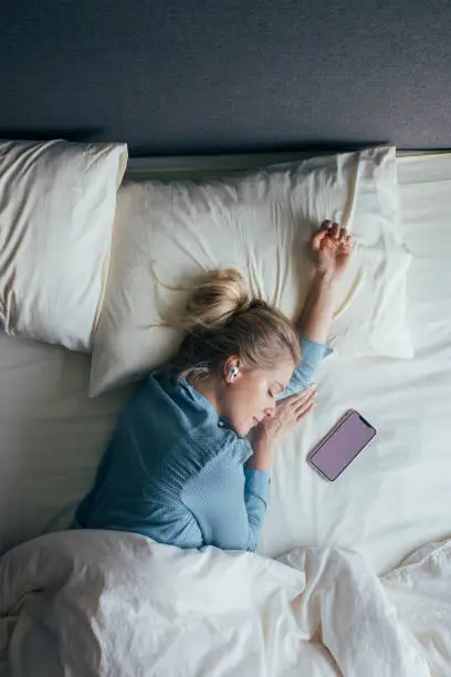 Photo of Technology-Aided Sleep: Woman in Pyjamas With Wireless Headphones Listening to Relaxing Music on Her Smartphone in Bed in the Morning