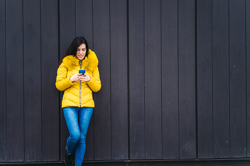 Cheerful young Caucasian woman with mobile phone, leaning on the wall.