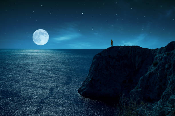 la persona è in piedi sul bordo di una scogliera contro il mare e la luna piena, sotto le stelle e la luce della luna. - phosphorescence foto e immagini stock