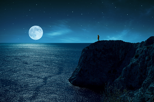 Person is standing in the edge of a cliff against the sea and full moon, under stars and moon light.