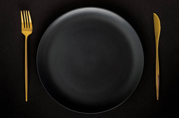 istock Black empty ceramic plate with golden cutlery (fork and knife) on a black background (table). 1211560535