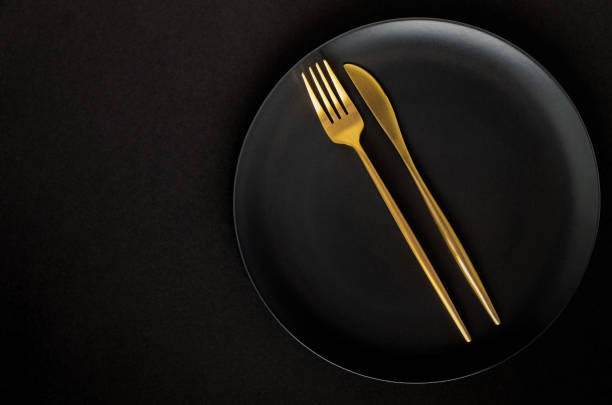 istock Black empty ceramic plate with golden cutlery (fork and knife) on a black background (table). 1211560555