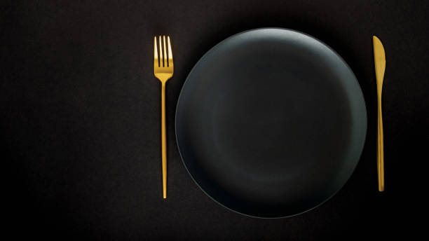 istock Black empty ceramic plate with golden cutlery (fork and knife) on a black background (table). 1211560615