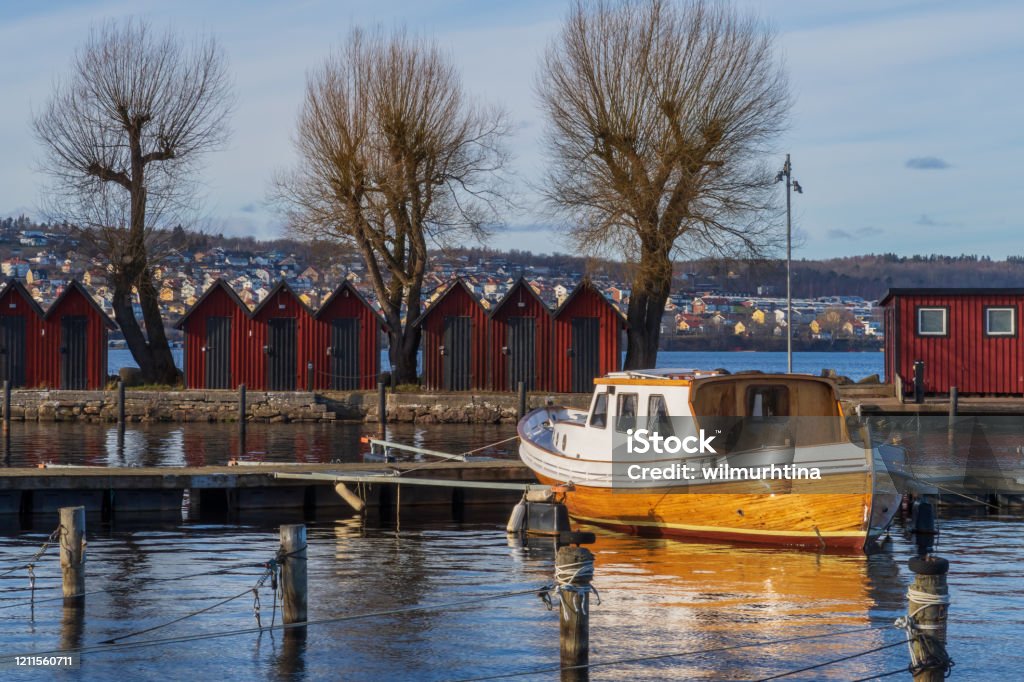 Boathouses and a small fishing boat in the harbor of Jönköping; Lake Vättern in the south of Sweden, a sunny day in february Traditional red boat houses in a row along the pier in Jönköpings harbor and an old fishing boat in the afternoon sun Harbor Stock Photo