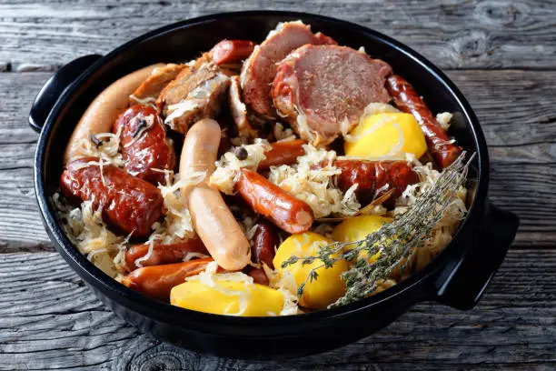 Alsace dish: sour cabbage stew with meat: pork loin and bacon and sausages and potato cooked in white wine thyme, juniper berries, garlic served on a black baking dish on wooden background, close-up