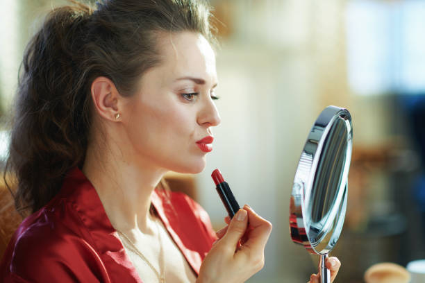 femme au foyer heureuse appliquant le rouge à lèvres rouge tout en regardant dans le miroir - wall indoors chair floor photos et images de collection