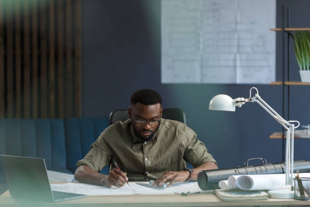 istock Afro-American architect working in office with blueprints.Engineer inspect architectural plan, sketching a construction project. Portrait of black handsome man sitting at workplace. Business concept 1211577091