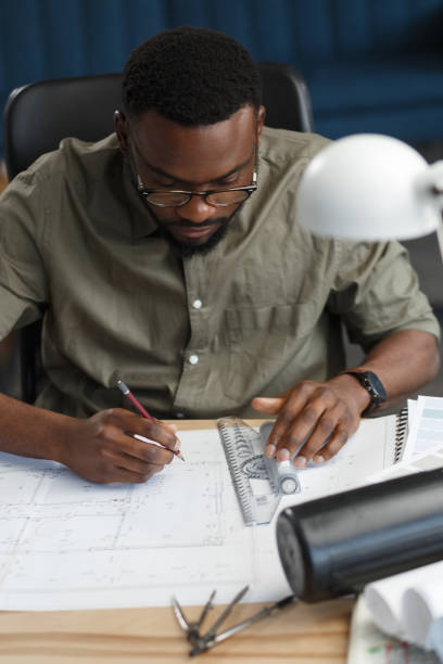 istock Afro-American architect working in office with blueprints.Engineer inspect architectural plan, sketching a construction project. Portrait of black handsome man sitting at workplace. Business concept 1211577659
