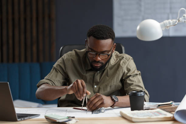 istock Afro-American architect working in office with blueprints.Engineer inspect architectural plan, sketching a construction project. Portrait of black handsome man sitting at workplace. Business concept 1211577675
