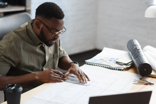 istock Afro-American architect working in office with blueprints.Engineer inspect architectural plan, sketching a construction project. Portrait of black handsome man sitting at workplace. Business concept 1211577795