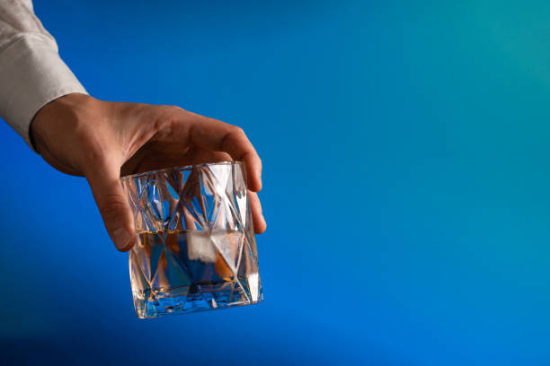 A human hand holds a glass of whiskey with ice on the blue background