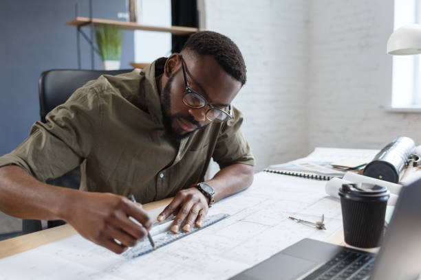 istock Afro-American architect working in office with blueprints.Engineer inspect architectural plan, sketching a construction project. Portrait of black handsome man sitting at workplace. Business concept 1211577952