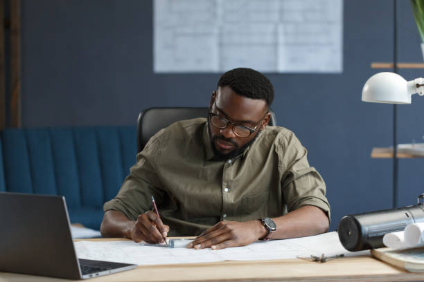 istock Afro-American architect working in office with blueprints.Engineer inspect architectural plan, sketching a construction project. Portrait of black handsome man sitting at workplace. Business concept 1211577989