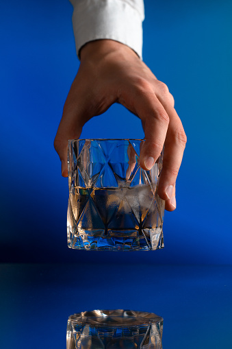 A human hand raises a glass of whiskey with ice on the blue background