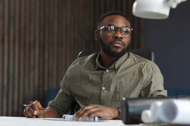 istock Afro-American architect working in office with blueprints.Engineer inspect architectural plan, sketching a construction project. Portrait of black handsome man sitting at workplace. Business concept 1211578011