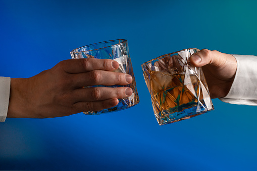 Close up of the hands of two people reaching to clink glasses of whiskey on the blue background
