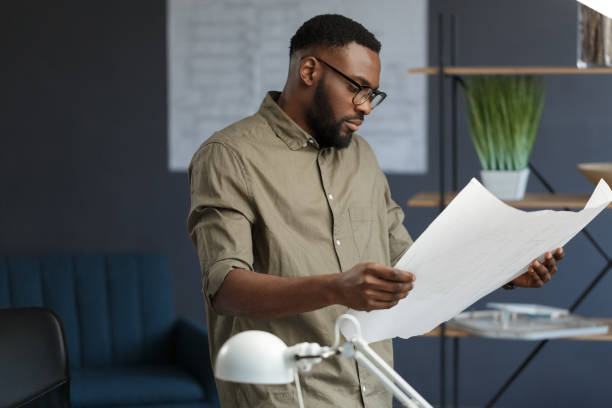 istock Afro-American architect working in office with blueprints.Engineer inspect architectural plan, sketching a construction project. Portrait of black handsome man sitting at workplace. Business concept 1211578082