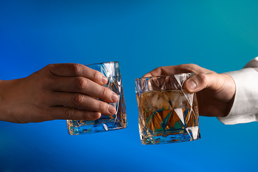 Close up of the hands of two people reaching to clink glasses of whiskey on the blue background