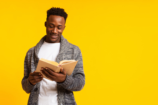 Romantic african guy reading poetry in old book on yellow background, copy space