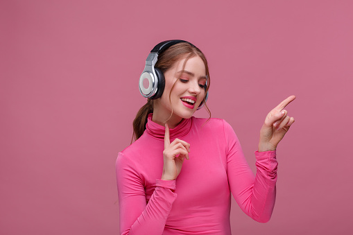 Enjoy listening to music. Beautiful young redhead woman in headphones listening music. Funny smiling girl in earphones and pink blouse dancing on pink background. Relaxation and stress management.