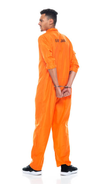 etnia africana joven hombre criminal de pie frente a fondo blanco usando uniforme y usando esposas - orange uniform fotografías e imágenes de stock