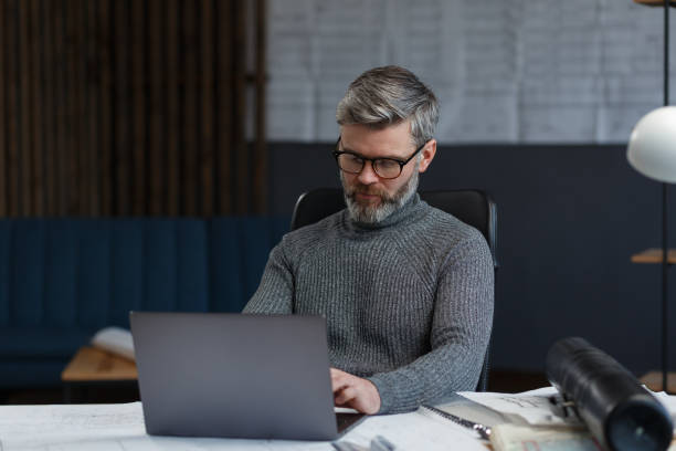 istock Designer working in office with laptop. Architect thinks over architectural plan, searching new ideas for construction project. Portrait of handsome bearded man sitting at workplace. Business concept 1211602977