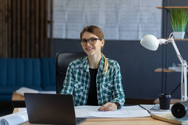 istock Female interior designer working in office with laptop. Architect thinks over architectural plan, searching new ideas for construction project. Woman sitting at workplace. Business portrait concept 1211605098
