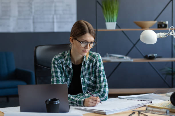 istock Female interior designer working in office with laptop. Architect thinks over architectural plan, searching new ideas for construction project. Woman sitting at workplace. Business portrait concept 1211605130