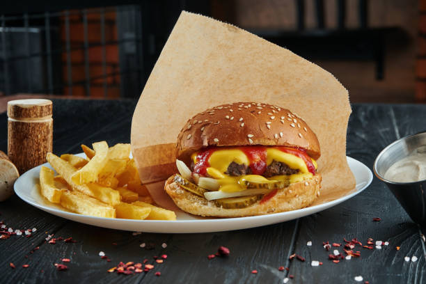 istock Appetizing and juicy burger with beef, melted cheddar cheese, ketchup, onions and pickled cucumbers on a white plate with french fries. Close up 1211606190