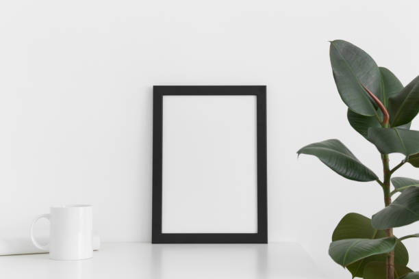 istock Black frame mockup with a mug, book and ficus on a white table. Portrait orientation. 1211616315