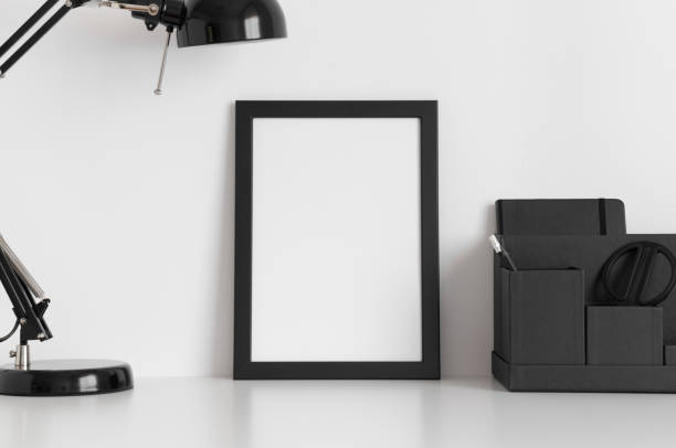 istock Black frame mockup with a lamp and workspace accessories on a white table. Portrait orientation. 1211616349