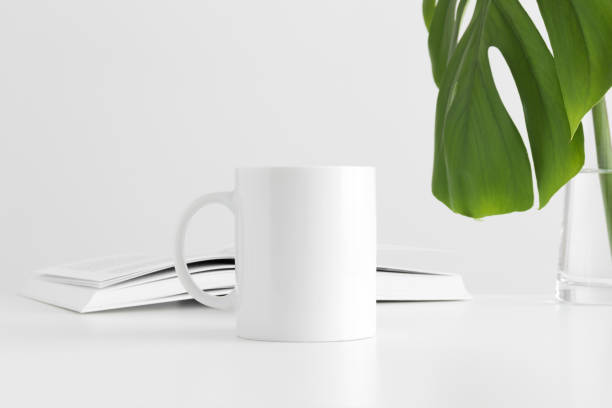 istock White mug mockup with a monstera leaf in a vase and a book on a white table. 1211616448