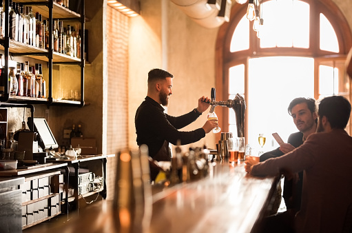 Shot of men sitting at bar counter with bartender filling beer glass from beer tap