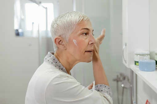 Portrait of Beautiful Senior Woman in Bathroom Applying Moisturizing Cream. Beauty, Skin Care and People Concept - Smiling Mature Woman Applying Cream to Face and Looking to Mirror at Home Bathroom. Healthy Skin Care Beauty Routine Treatment Concept
