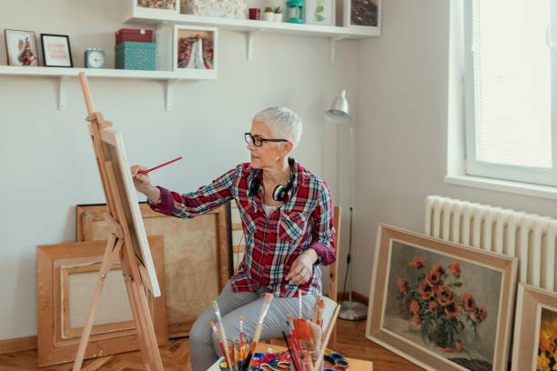 60-year-old Latin woman with short white hair is sitting on a chair in the backyard of her house wearing a yellow apron doing her favorite hobby painting a beautiful picture on canvas