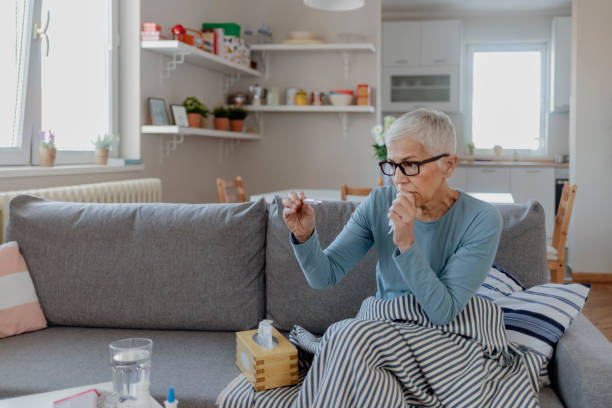 Sick Woman with a Cold Measuring Temperature Sick Senior Woman Sitting on Sofa Measuring Body Temperature With Thermometer Under the Blanket. Mature Woman Caught Cold or Flu Illness. Portrait of Unhealthy Woman With Seasonal Infections, Flu, Allergy Sitting in Bed in the Livingroom. High Temperature Concept. one mature woman only stock pictures, royalty-free photos & images