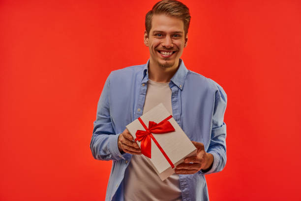 istock Happy guy with a beard in a blue shirt holds a present in a white box in hands. 1211640363