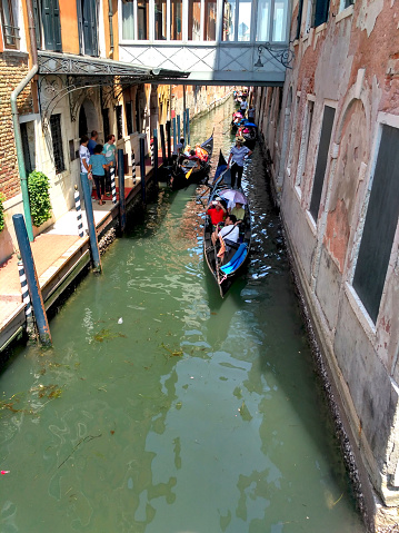 Gondolas with Asian and European tourists pass through water canal in Venice. Venecian boats, tourists, water canal between buildings. People watching gondolas.