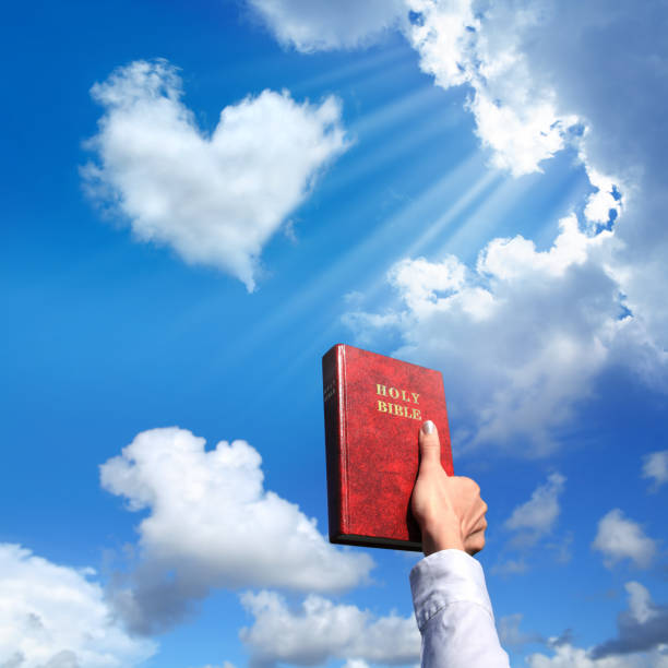 Female hand holding Holy Bible over shiny blue sky with heart shaped cloud