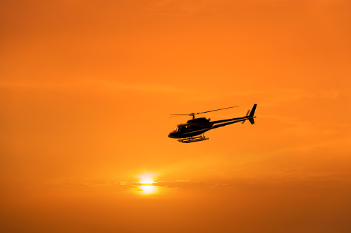 Silhouetted one private helicopter flying over orange color sunset sky